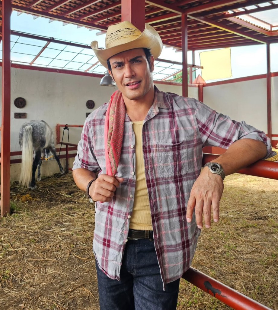Actor posando en un establo, vistiendo una camisa a cuadros, un sombrero de vaquero y rodeado de caballos.