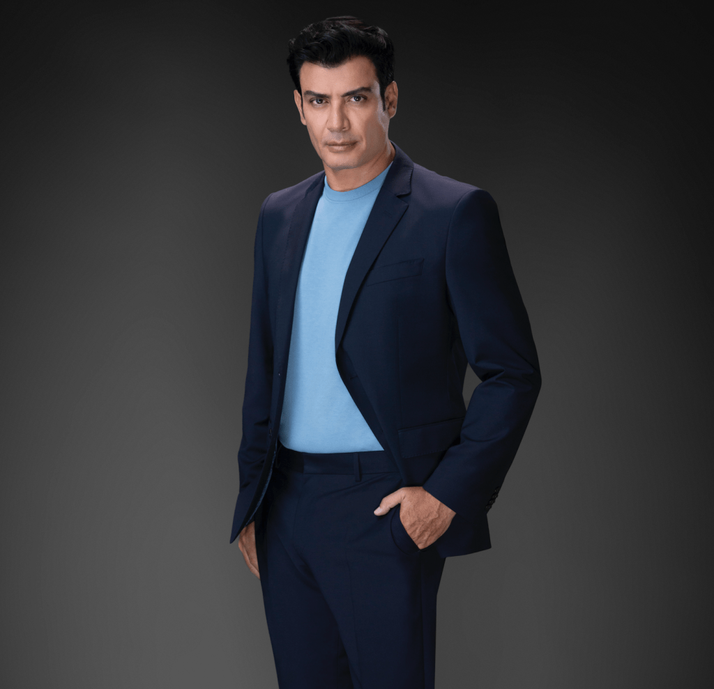 Actor posando en un fondo gris oscuro, vistiendo un traje en tonos oscuros y una camiseta azul clara.