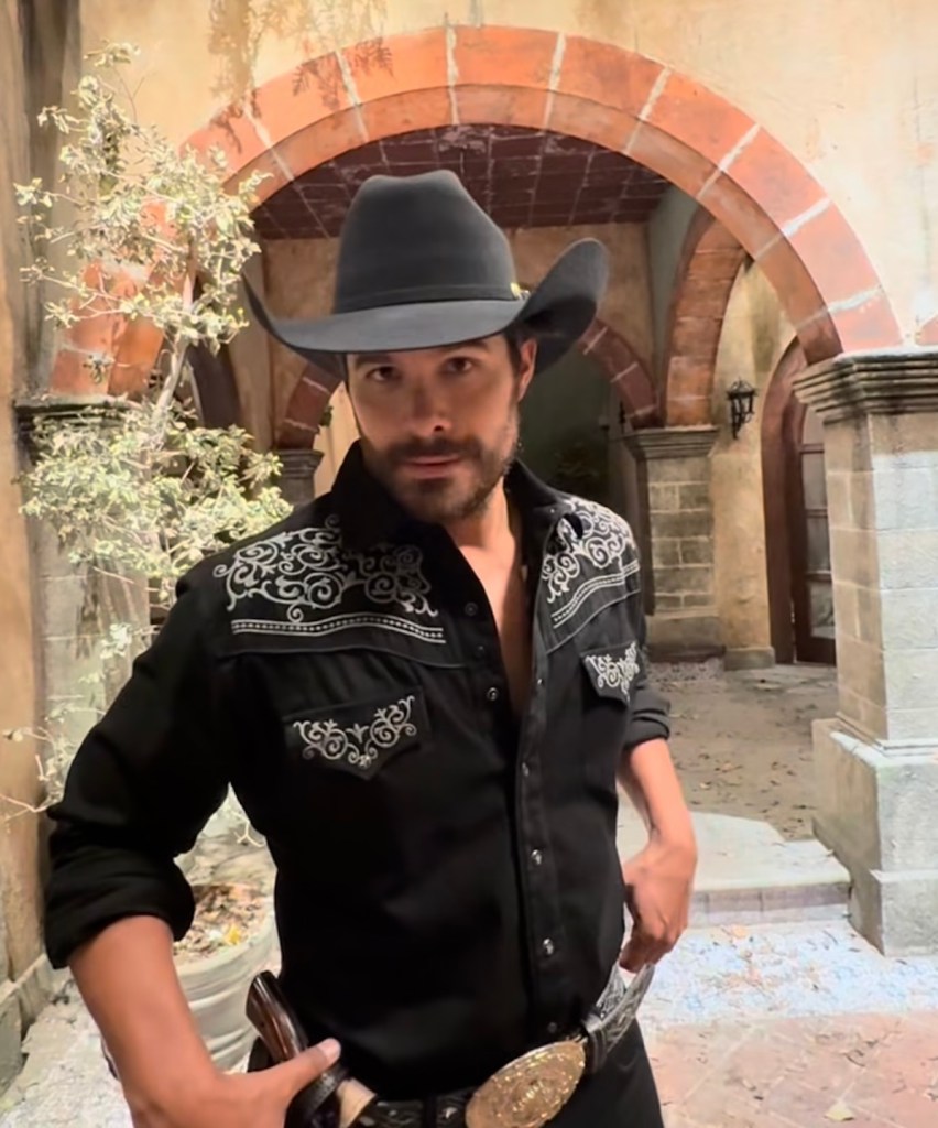 Hombre posando con sombrero negro y camisa negra decorada, en un entorno arquitectónico antiguo.