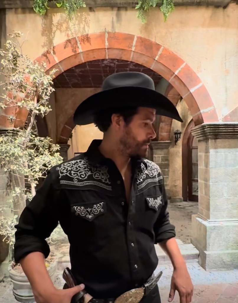 Hombre con sombrero de charro y camisa negra decorada, posando en un escenario con elementos arquitectónicos coloniales.