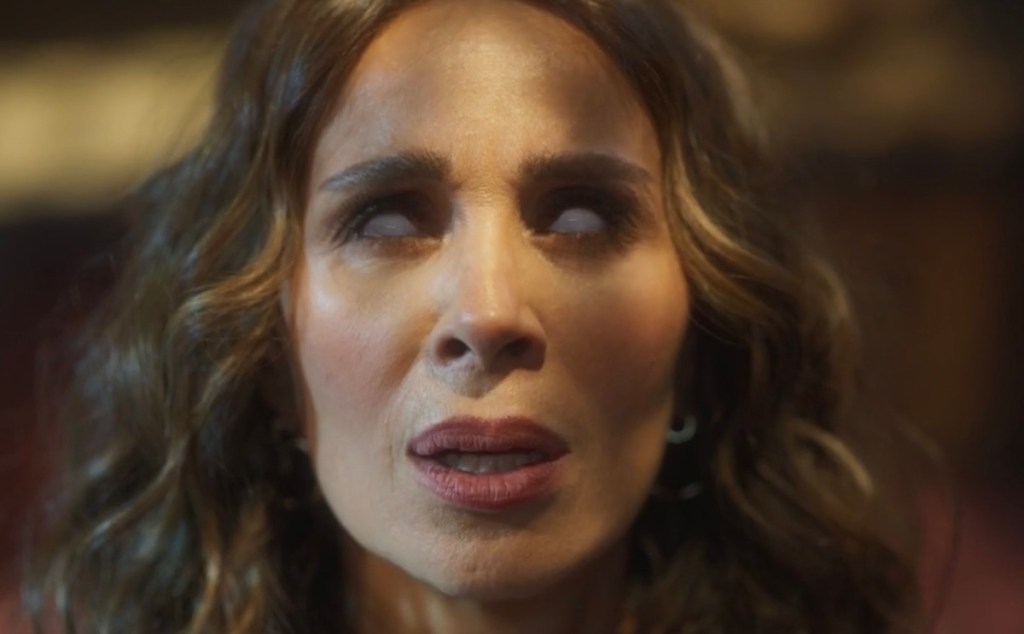 Close-up of a woman with curly hair, looking up with white eyes and an intense expression, portraying an eerie character in a dramatic scene.