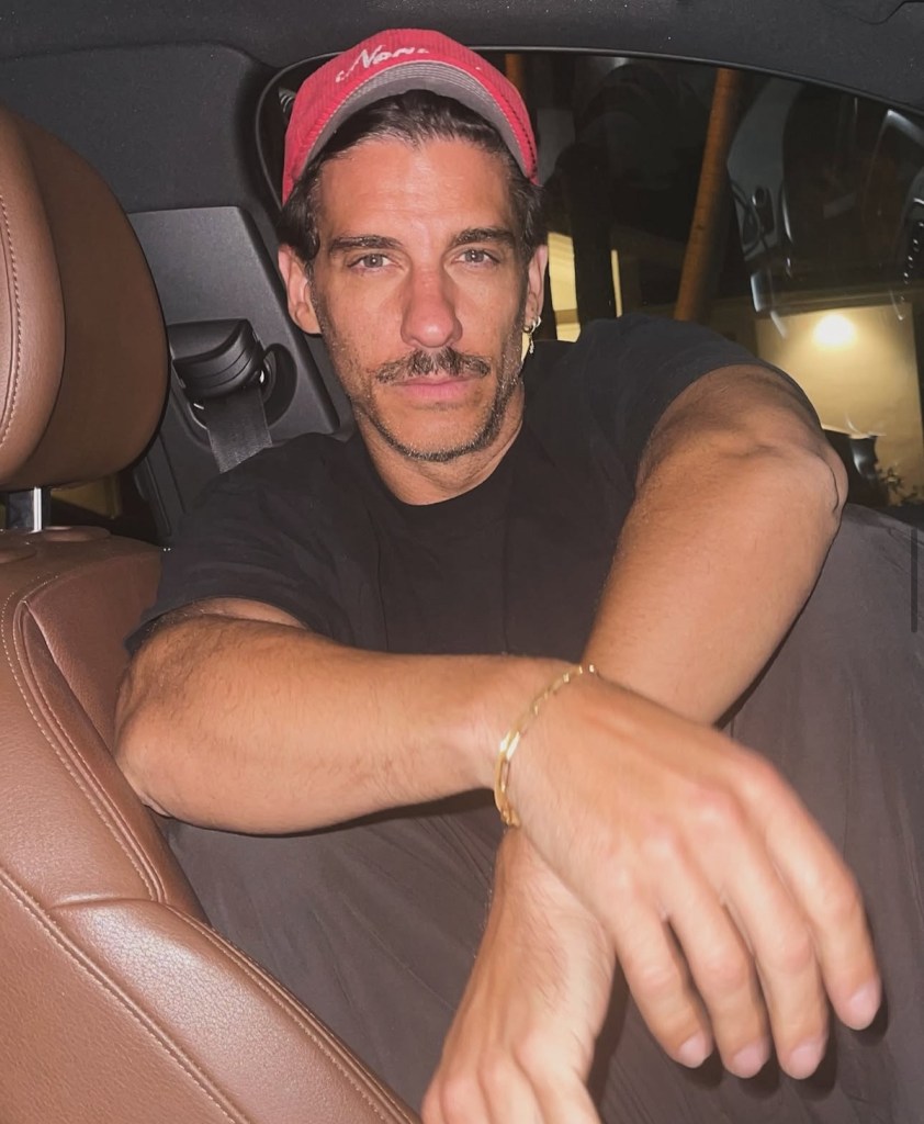 Retrato de un hombre sentado en el interior de un coche, con una gorra roja y una camiseta negra, mostrando una postura relajada.