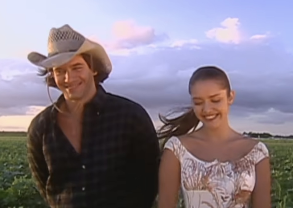 Escena de la telenovela _Gata salvaje_, con dos personajes sonrientes, uno usando un sombrero y el otro con un vestido floral, de pie en un campo al atardecer.