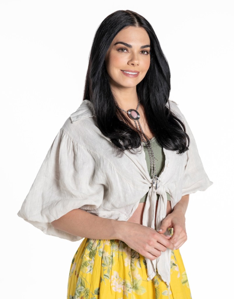 Mujer sonriente con cabello largo y oscuro, vestida con una blusa clara y una falda amarilla con flores, posando con las manos juntas.
