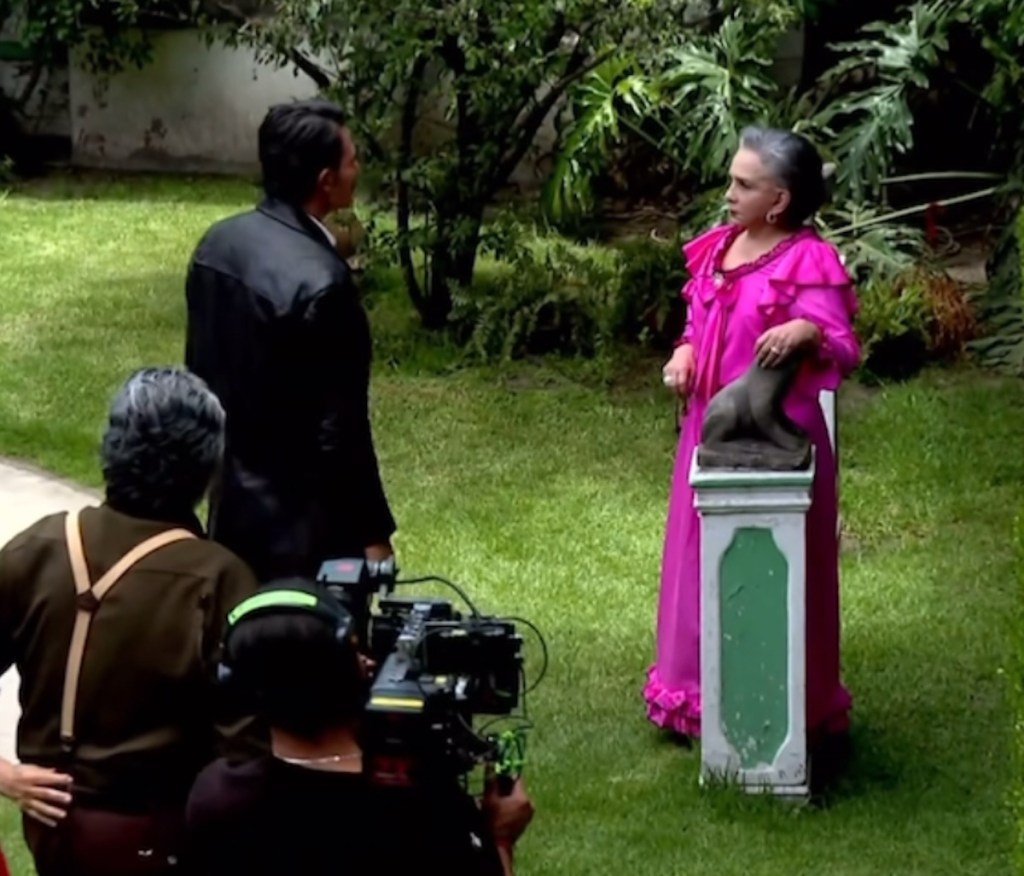 Escena del rodaje de la telenovela Amanecer, mostrando a una actriz con vestido rosa y un actor de espaldas frente a una estatua en un jardín.