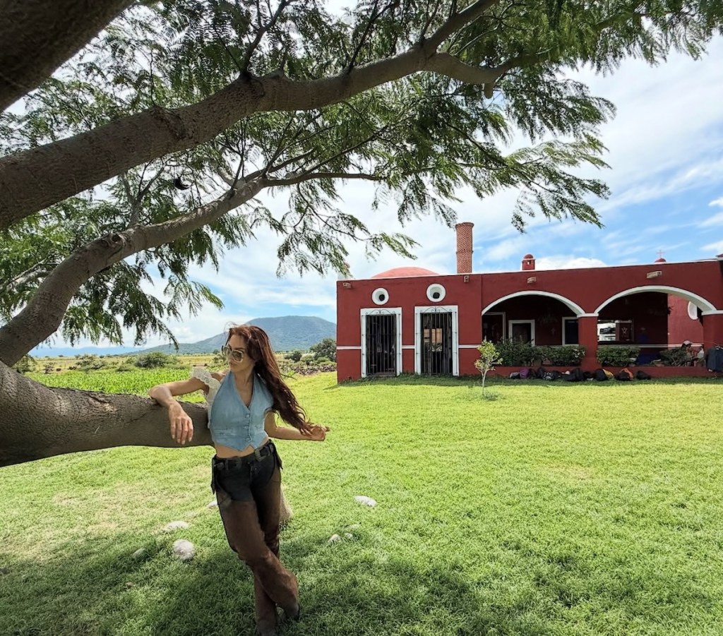 Mujer apoyada en un árbol, luciendo un top de mezclilla y pantalones oscuros, frente a una casa roja en un paisaje natural.