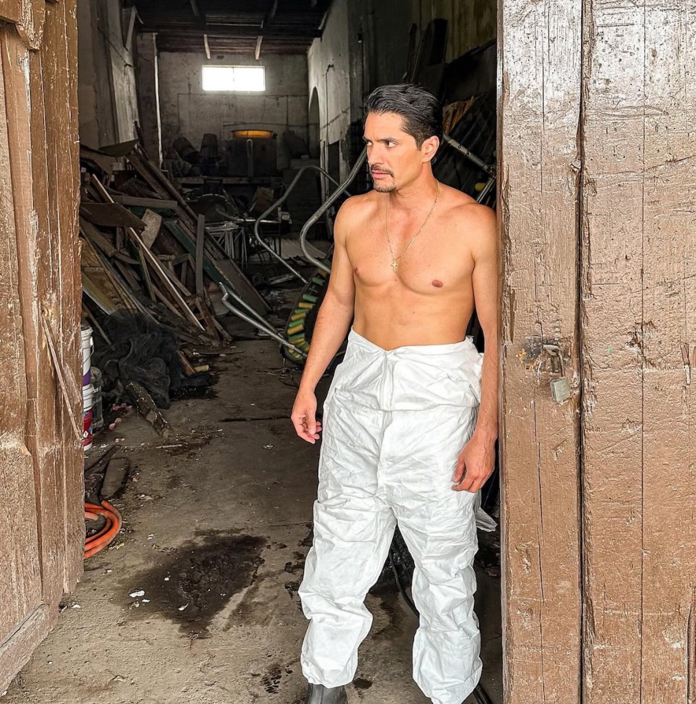 Hombre con pantalones blancos y sin camiseta en un espacio desordenado, mirando hacia el frente.