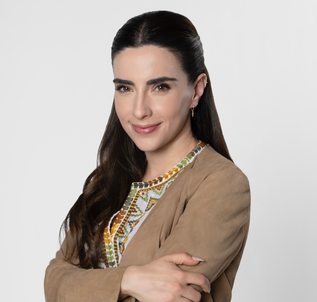 Retrato de una mujer sonriente con pelo largo y lacio, vestida con una prenda de color beige y decorada con un diseño colorido. Ella cruza los brazos y se presenta sobre un fondo blanco.