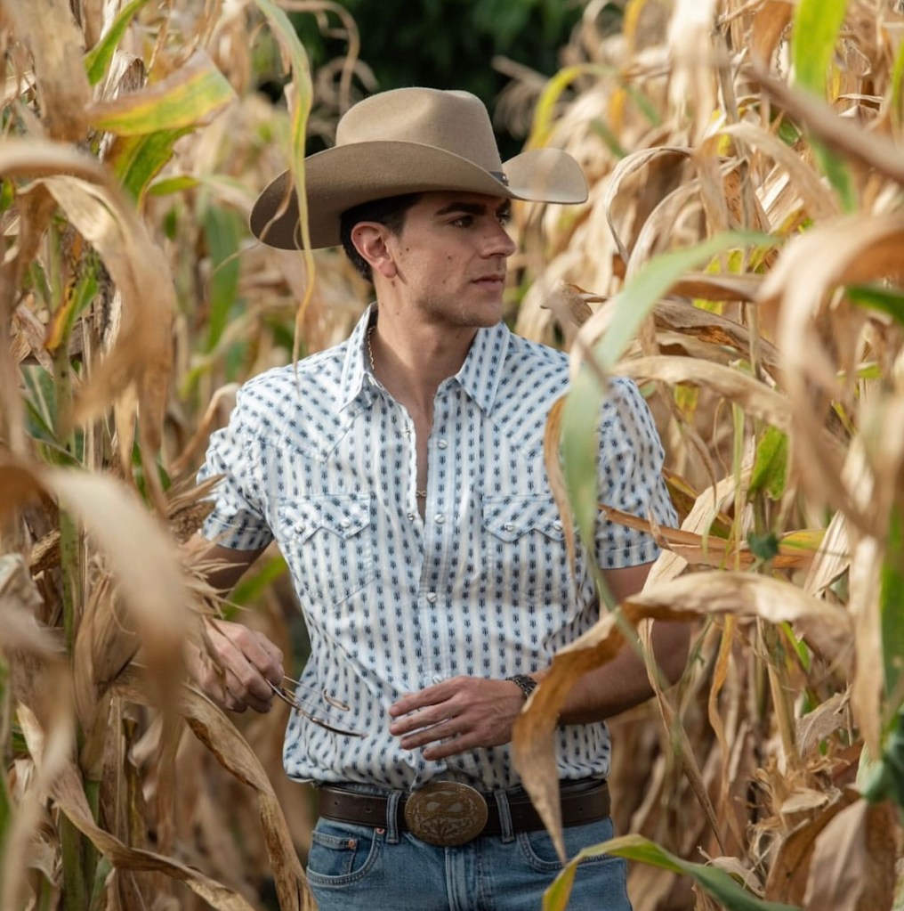 Hombre joven con camisa de cuadros y sombrero, posando entre maizales secos.