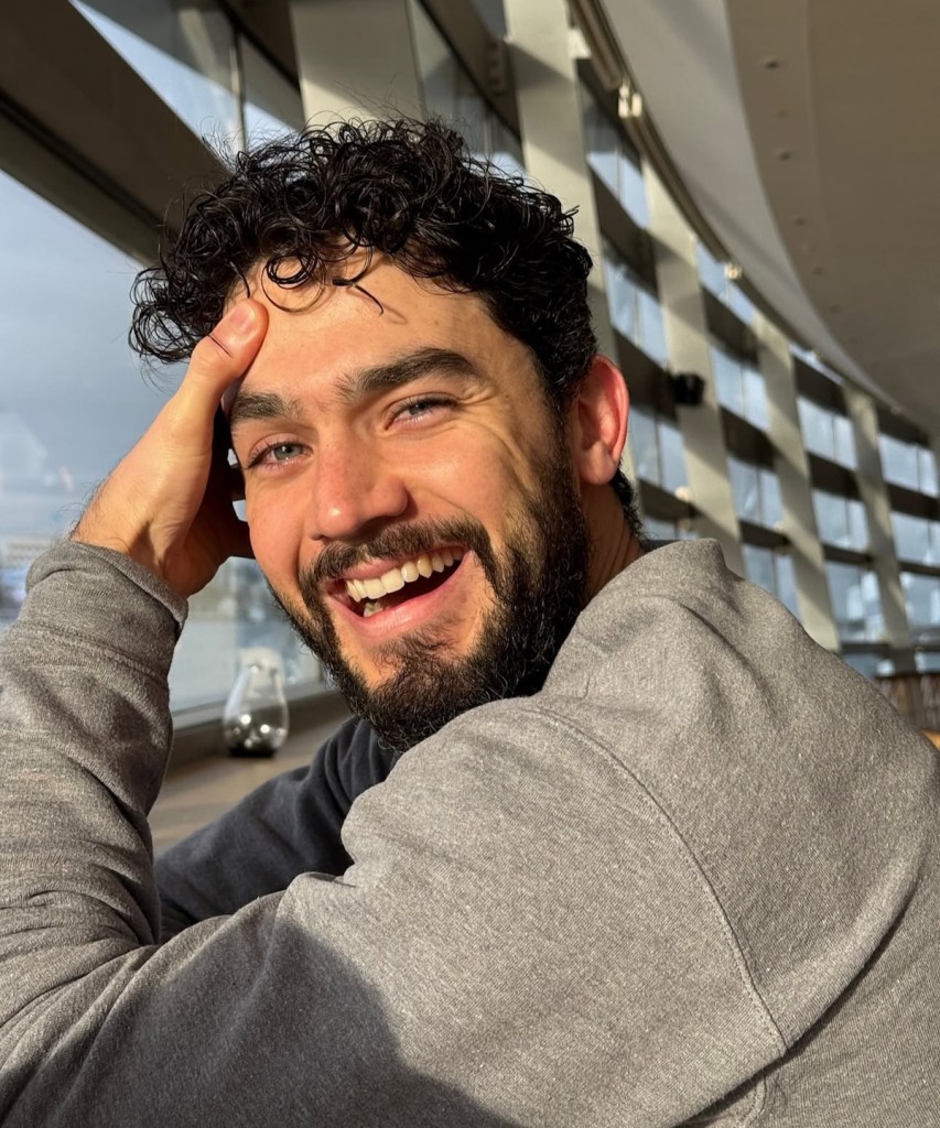 Un hombre sonriendo con barba y cabello rizado, sentado junto a una ventana luminosa.