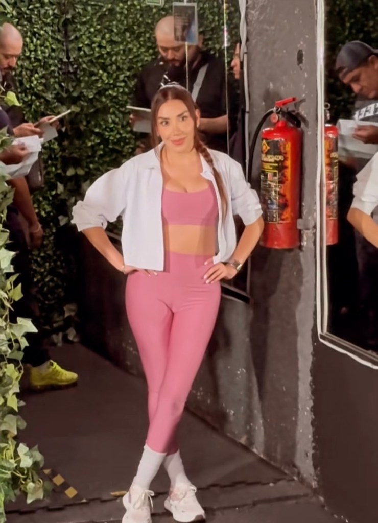 Karime Pindter posando en un set de grabación, vistiendo un top rosa y leggings a juego, con una chaqueta blanca y zapatillas deportivas. En el fondo, se puede ver a varias personas trabajando en el set.