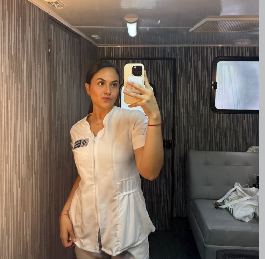Karla Esquivel tomando un selfie en un vestidor, vistiendo un uniforme blanco, con un fondo de pared decorada y un sofá al lado.