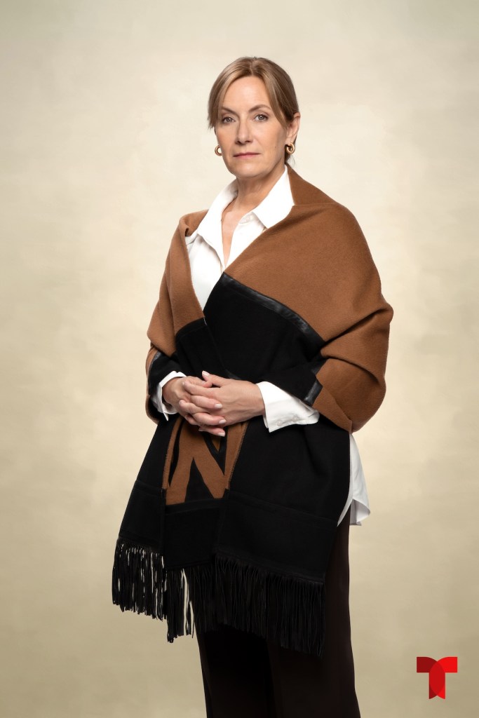 Retrato de una mujer con cabello rubio y un chal en tonos marrón y negro, cruzando los brazos frente a un fondo neutro.