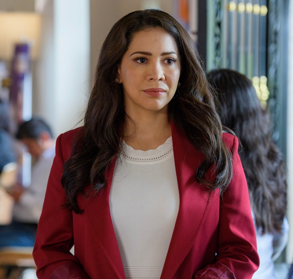 Mujer con cabello largo y suelto, viste una blusa blanca y un blazer rojo, mirando al frente con una expresión seria en un entorno interior.