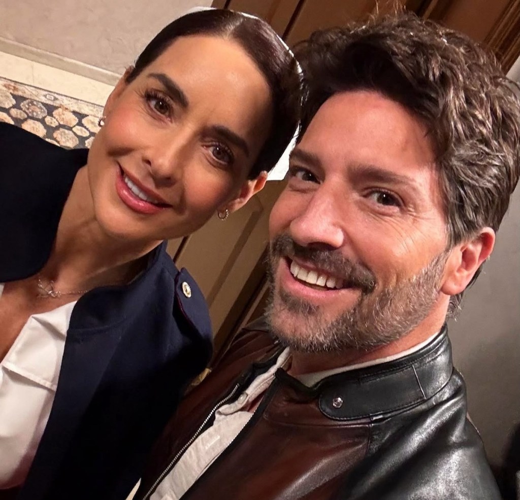 Selfie de dos actores sonrientes, uno con cabello corto y barba, y la otra con cabello recogido. Ambos están en un entorno interior con fondo desenfocado.
