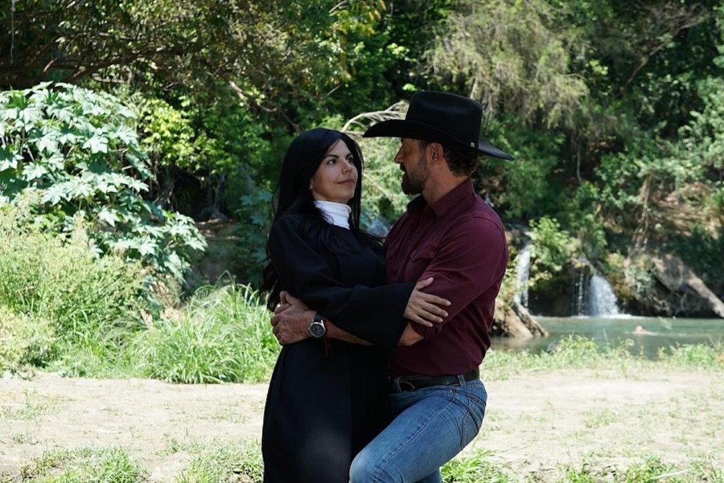 Una mujer vestida de monja abraza a un hombre con sombrero en un entorno natural con vegetación y agua de fondo.