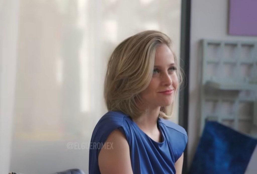 Mujer con cabello rubio y suelto, vestida con una blusa azul, sonriendo mientras se sienta en un espacio moderno con fondo difuso.