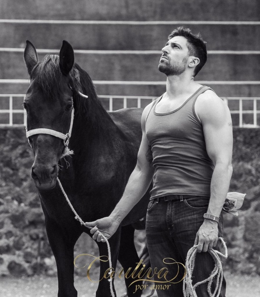 Hombre de pie junto a un caballo negro, luciendo una camiseta sin mangas, mirando hacia arriba en un entorno al aire libre.
