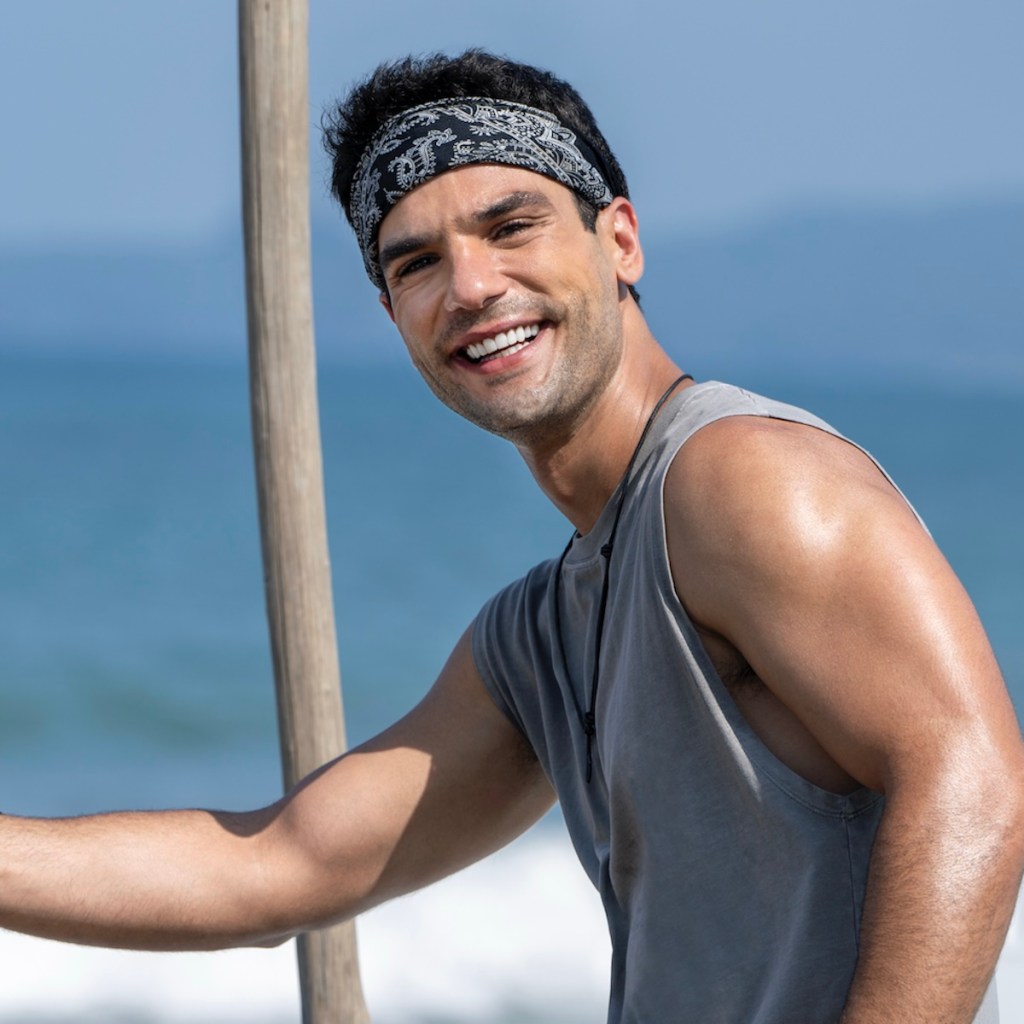 Hombre sonriendo en una playa, usando una camiseta sin mangas y un pañuelo en la cabeza, con el mar de fondo.