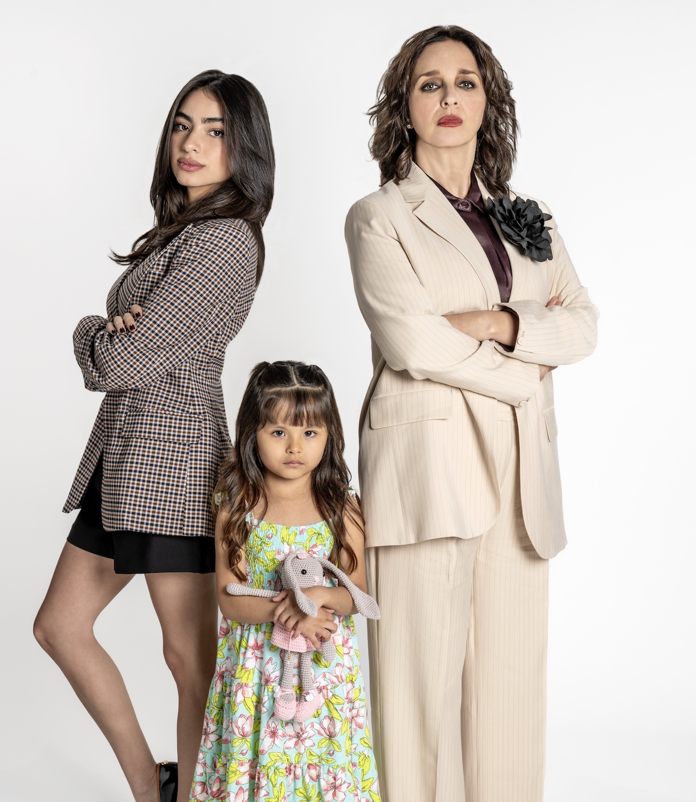 Isabella, Eugenio y Valentina posan juntos, la primera viste un abrigo con estampado, Valentina lleva un vestido colorido y sostiene un muñeco, mientras que la tercera usa un traje claro y tiene una expresión seria.