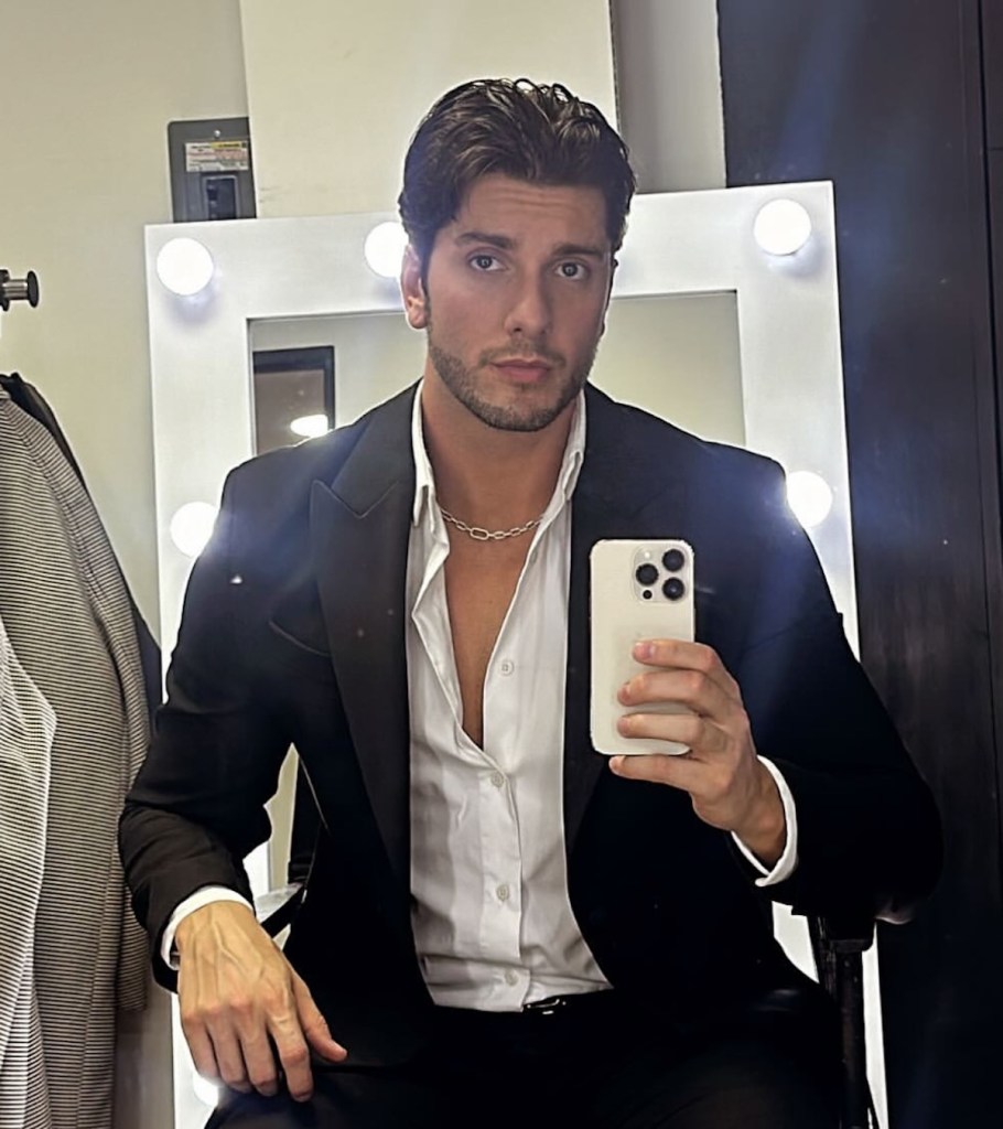 Selfie de un hombre con cabello ondulado y bien cuidado, usando un blazer negro y una camisa blanca. El fondo muestra un espejo con luces alrededor, creando un ambiente de maquillaje o vestuario.