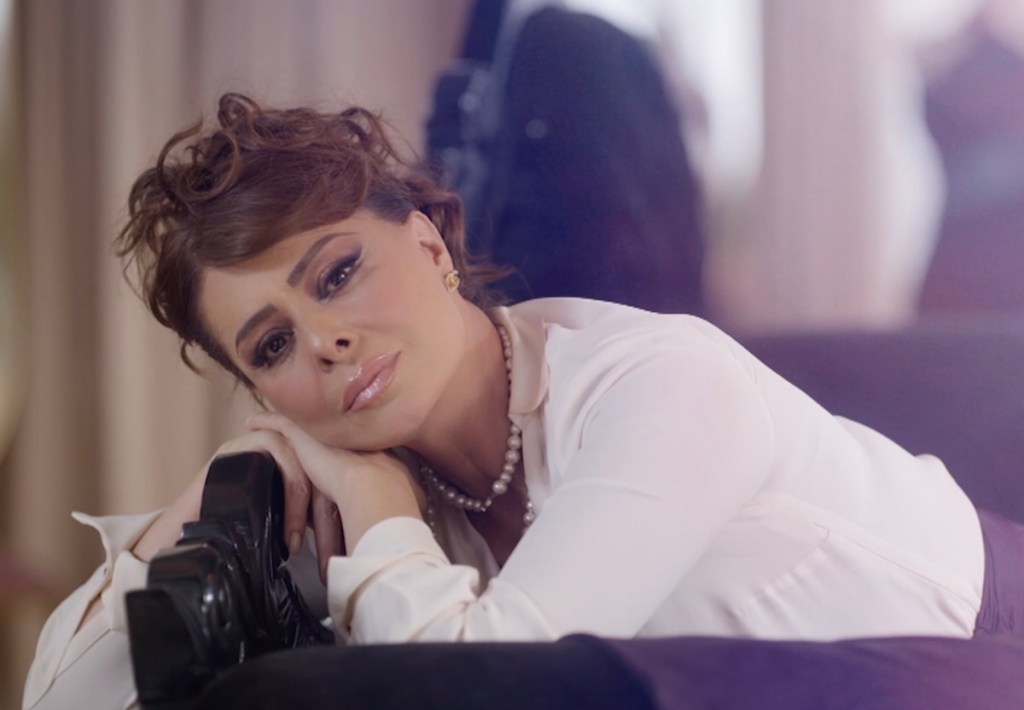 Mujer con cabello rizado y maquillaje pronunciado, acostada sobre un sofá, con una expresión melancólica y un collar de perlas, en un ambiente elegante.