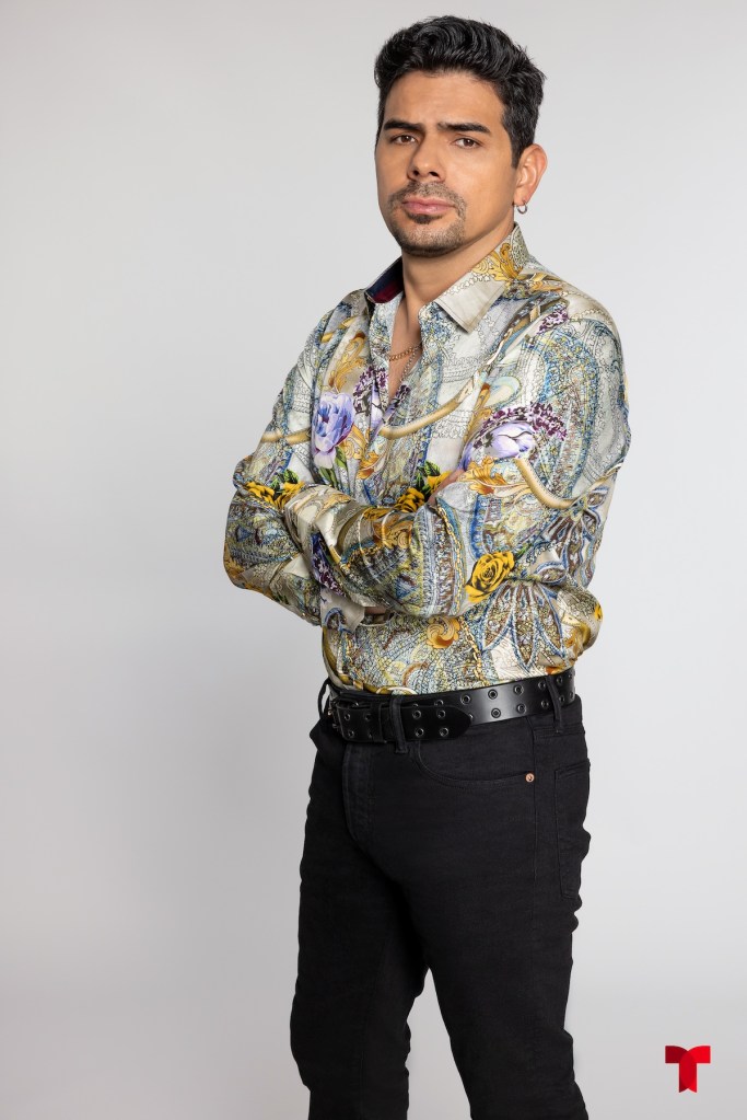 Hombre de pie con camisa colorida de estampado floral y pantalones oscuros, con una expresión seria y los brazos cruzados.