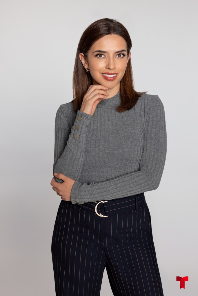 Mujer sonriendo, con cabello largo y castaño, vistiendo un suéter gris y pantalones de rayas, posando con una mano en el rostro.