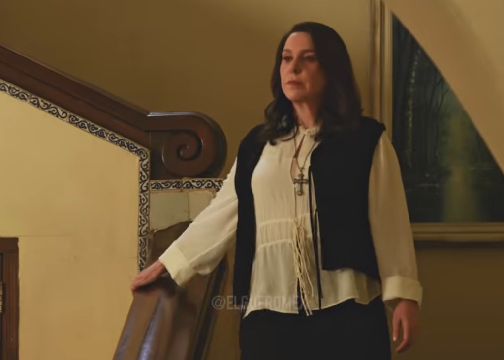 Una mujer con una blusa blanca y un chaleco negro, de pie frente a una escalera con una expresión seria en su rostro.