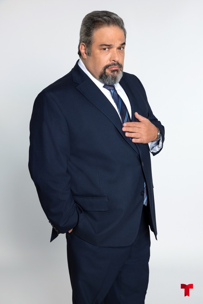 Hombre de mediana edad con barba y cabello canoso, vestido con un traje oscuro y corbata, posando con una mano sobre su pecho en un fondo blanco.