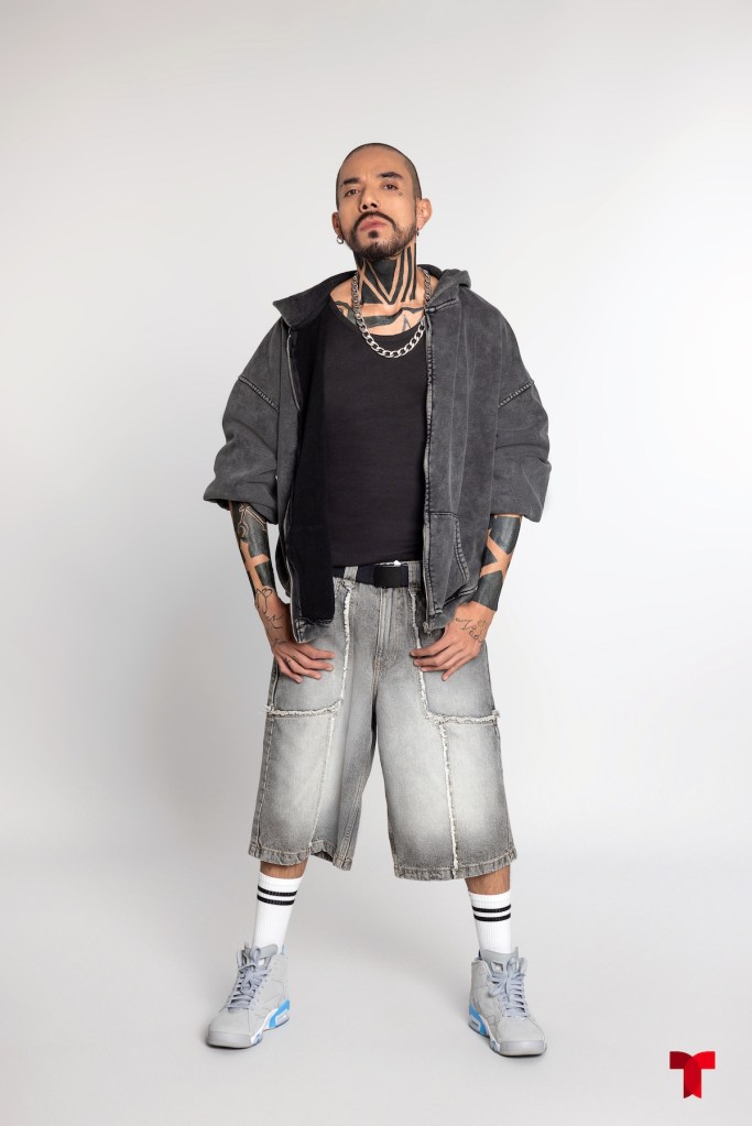 Hombre con tatuajes y cabello corto, vistiendo una camiseta negra y pantalones cortos de mezclilla, con cadenas y zapatillas deportivas en un fondo blanco.