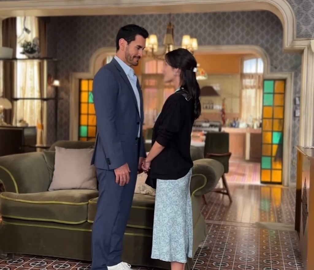 Escena de la telenovela 'Somos familia' con Oka Giner y David Zepeda en un ambiente acogedor de sala, ambos personajes interactuando.