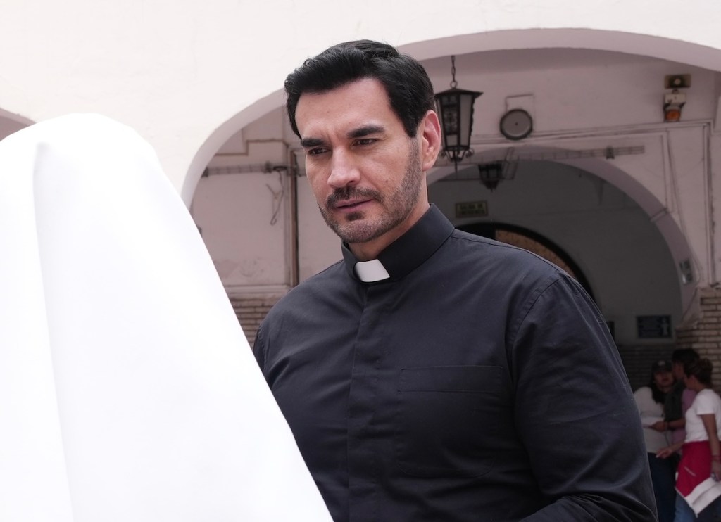 Un hombre con un collar de sacerdote observa con preocupación mientras sostiene una tela blanca, en un entorno arquitectónico tradicional.