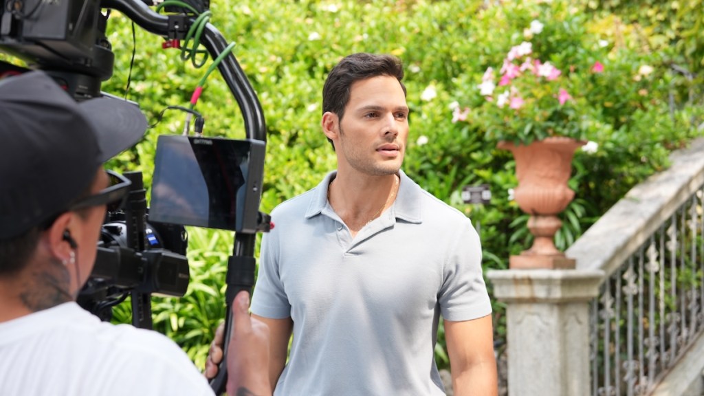 Un actor de pie en un set de filmación, frente a una cámara, rodeado de un jardín verde con flores.