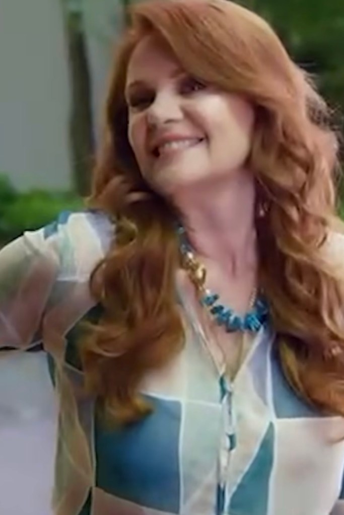 Mujer con cabello largo y rizado, sonriendo, vestida con una blusa de patrones en colores claros y un collar azul, de pie en un entorno exterior.