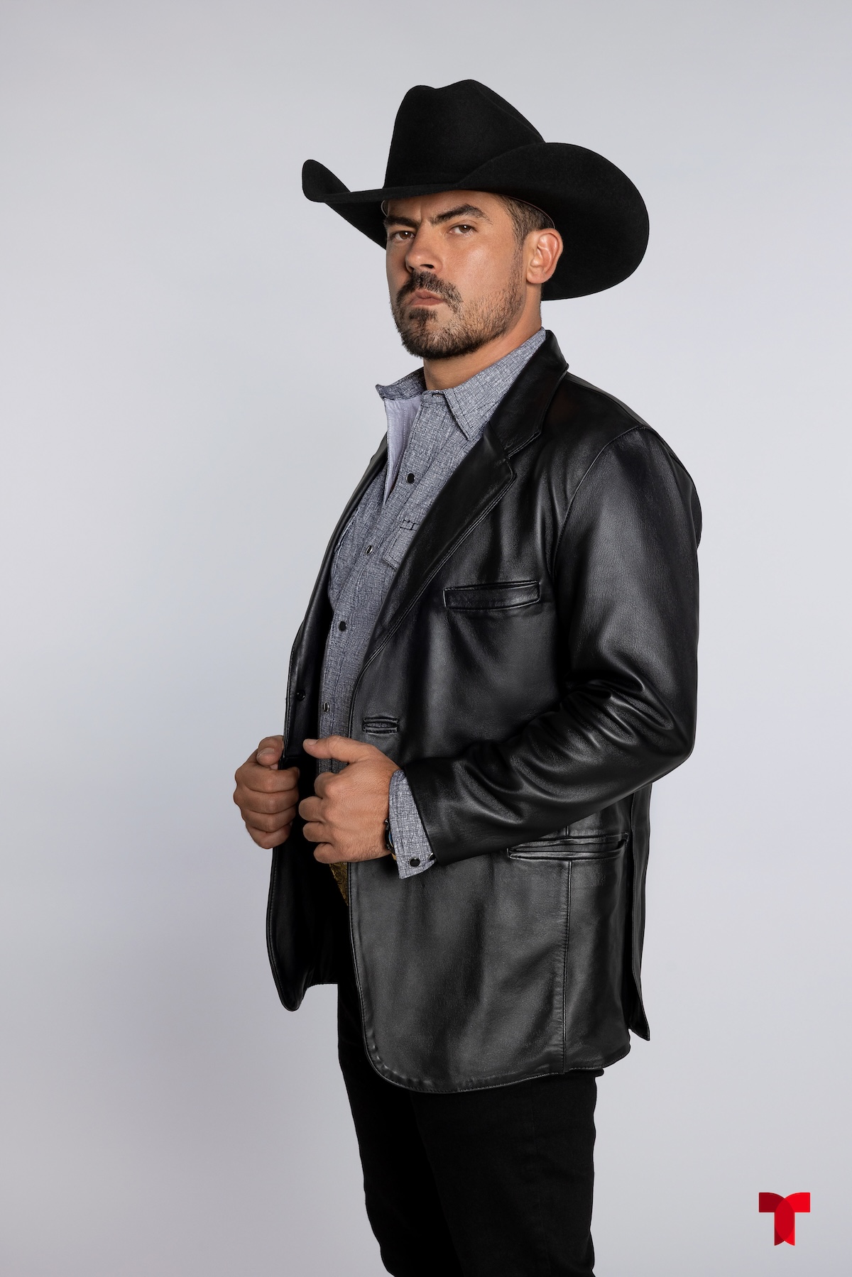 Hombre de pie con chaqueta de cuero negra y sombrero de vaquero, mirando hacia la cámara con una expresión seria.