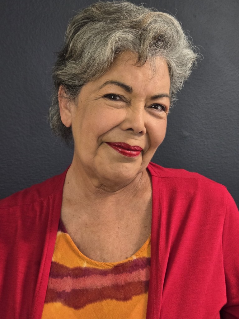 Retrato de Jeannette Lehr sonriendo, con cabello corto y canoso, vistiendo una blusa amarilla con rayas y una chaqueta roja. El fondo es de color azul oscuro.