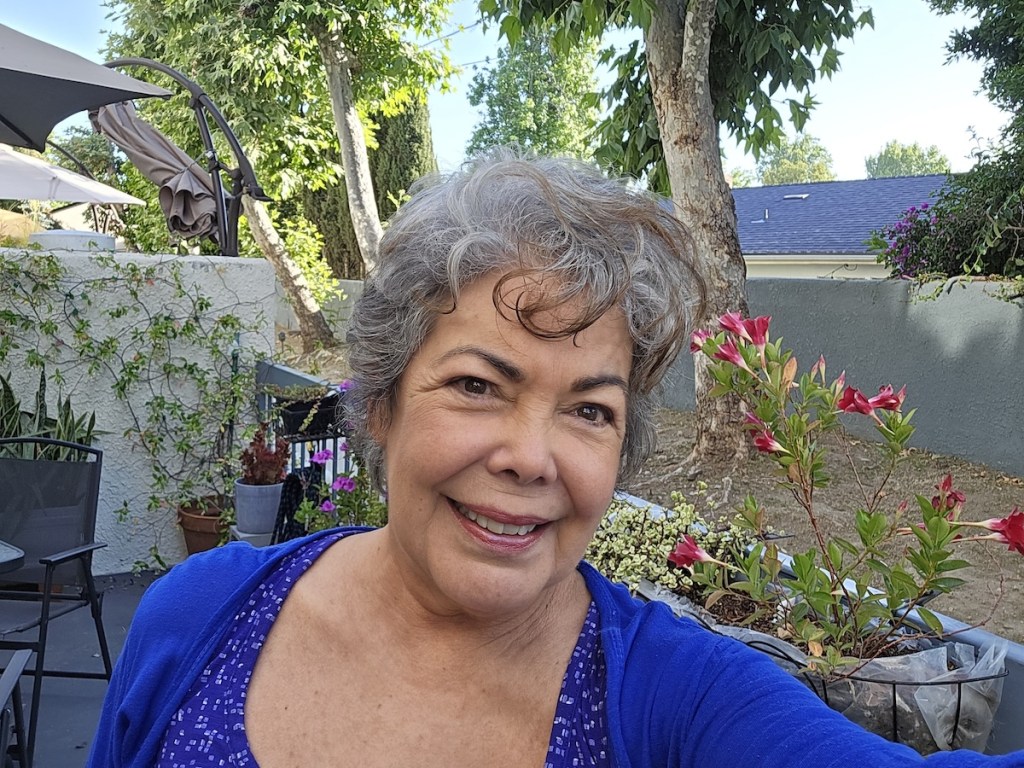 Selfie of Jeannette Lehr smiling in a garden setting with green plants and flowers around her.