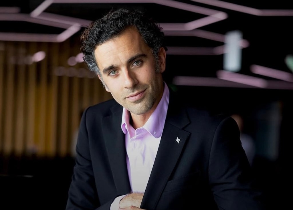 Retrato de un hombre con cabello rizado y una camisa de vestir rosa, sonriendo mientras se apoya con una mano en su pecho, en un ambiente moderno con luces suaves de fondo.
