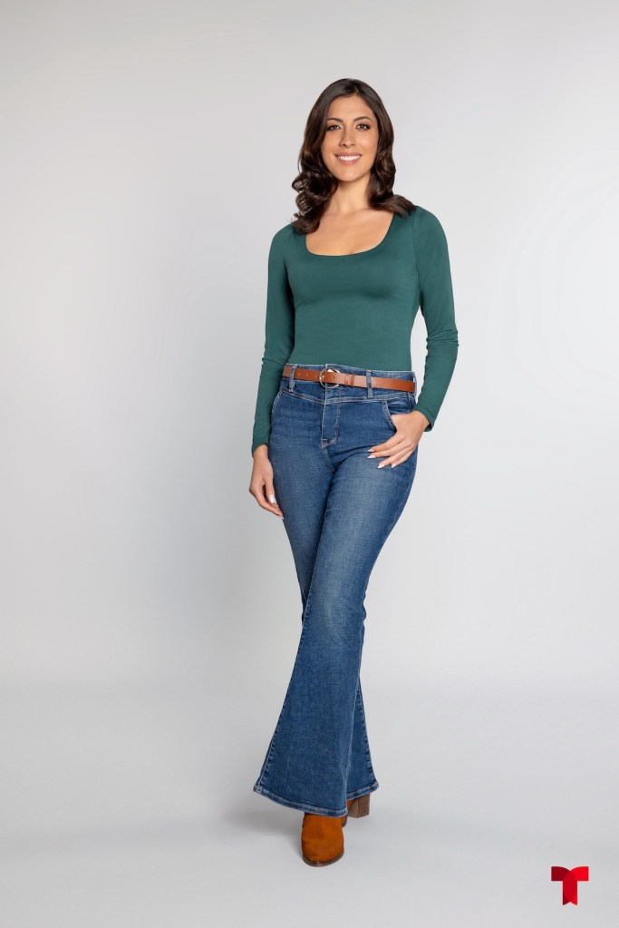 Mujer de pie sonriendo, llevando una camiseta verde y jeans acampanados, con una mirada amigable y un fondo claro.