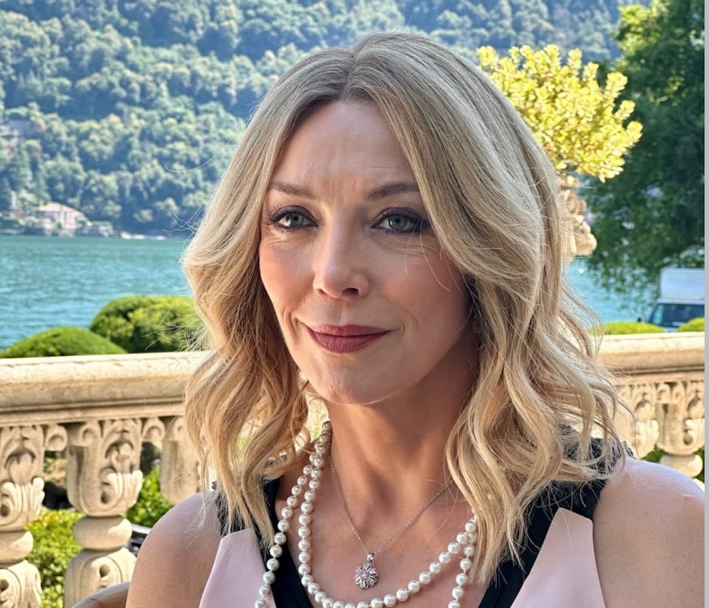 Mujer con cabello rubio y ondulado, vestida con un top negro y rosa, sonríe suavemente frente a un paisaje natural con un lago y montañas al fondo.