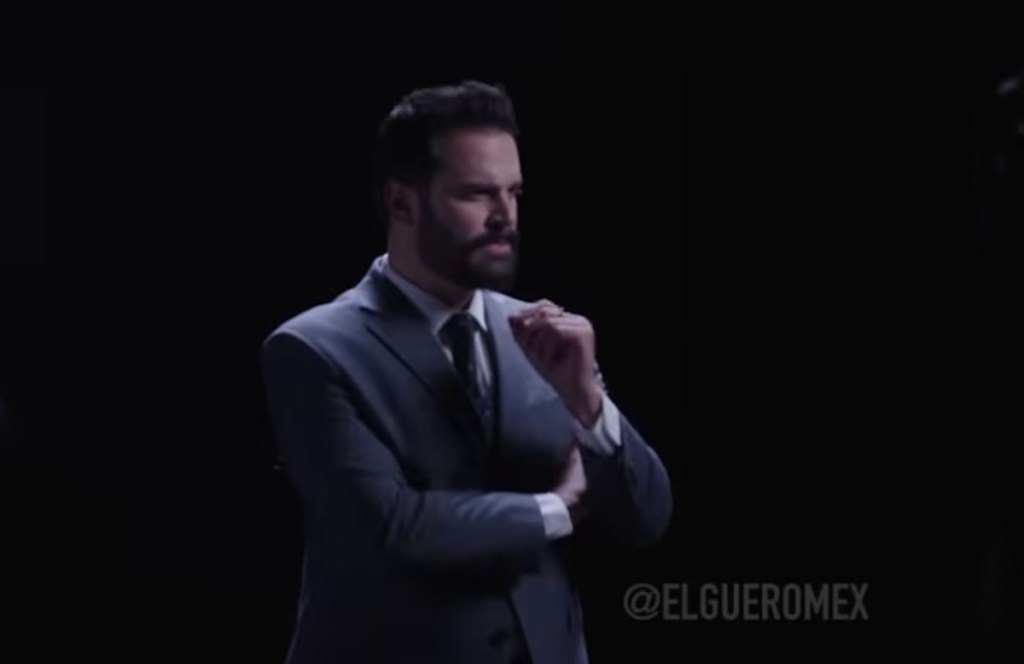 Hombre con traje en un fondo oscuro, mostrando una expresión contemplativa y gesticulando con la mano.
