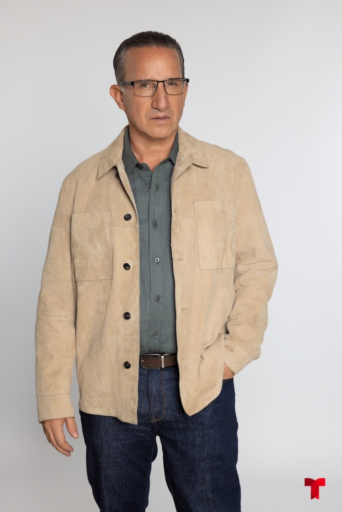Hombre de mediana edad con cabello corto y gafas, vistiendo una chaqueta de gamuza clara sobre una camisa de color gris y jeans oscuros, en un fondo blanco.