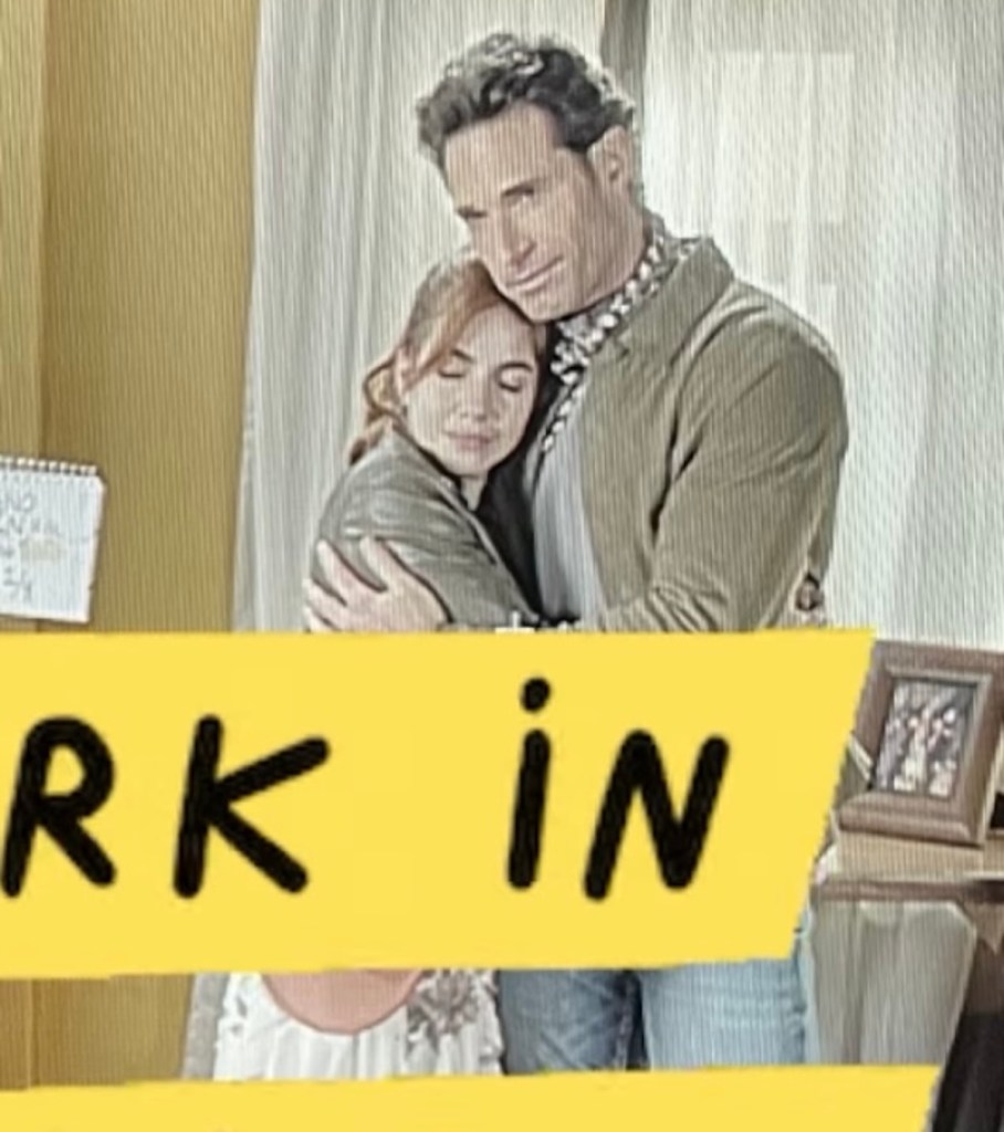 Un hombre y una mujer abrazados en un ambiente acogedor, con una decoración sencilla y un letrero amarillo que dice 'WORK IN'.