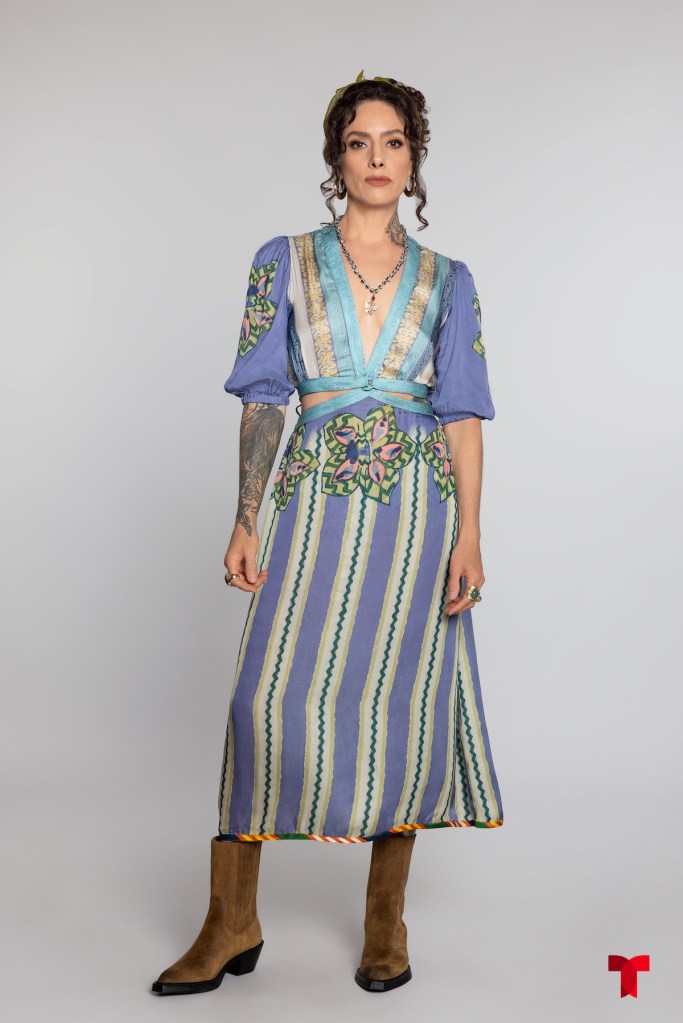 Una mujer con cabello rizado y labios pintados, vistiendo un vestido largo de colores azul y multicolor, con detalles florales y botas marrones, posando frente a un fondo gris claro.