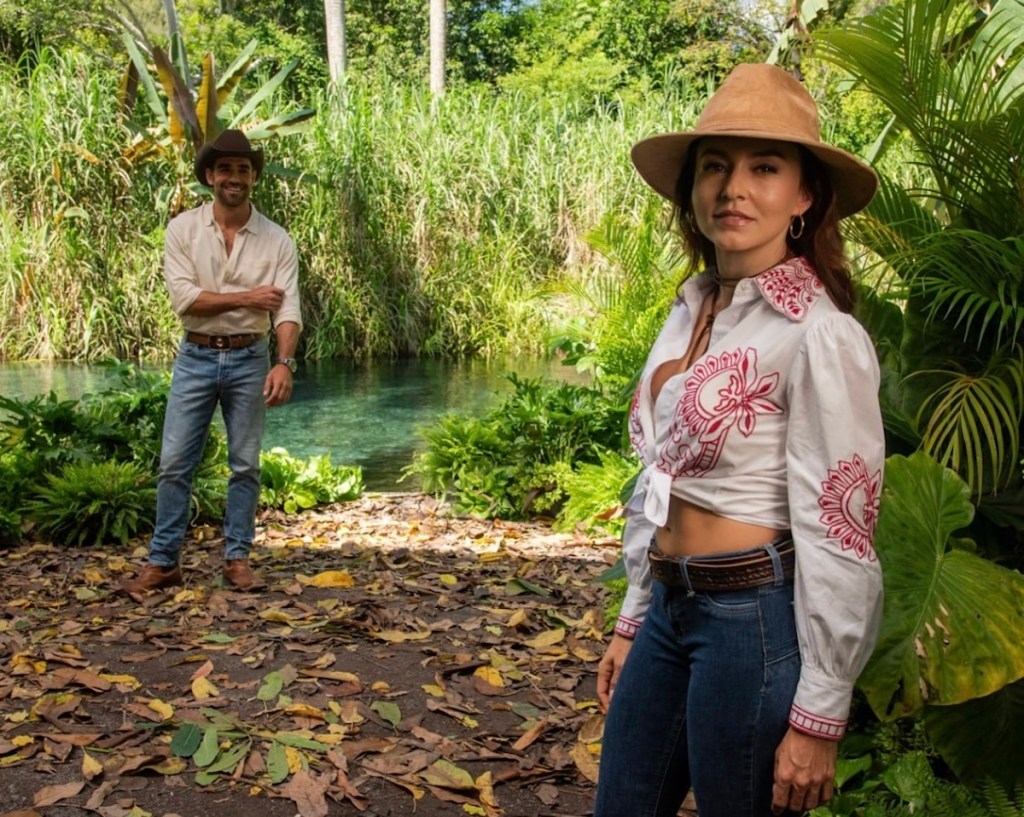 Una mujer con camisa blanca bordada y jeans posando frente a un hombre en un entorno natural con vegetación exuberante y un estanque al fondo.