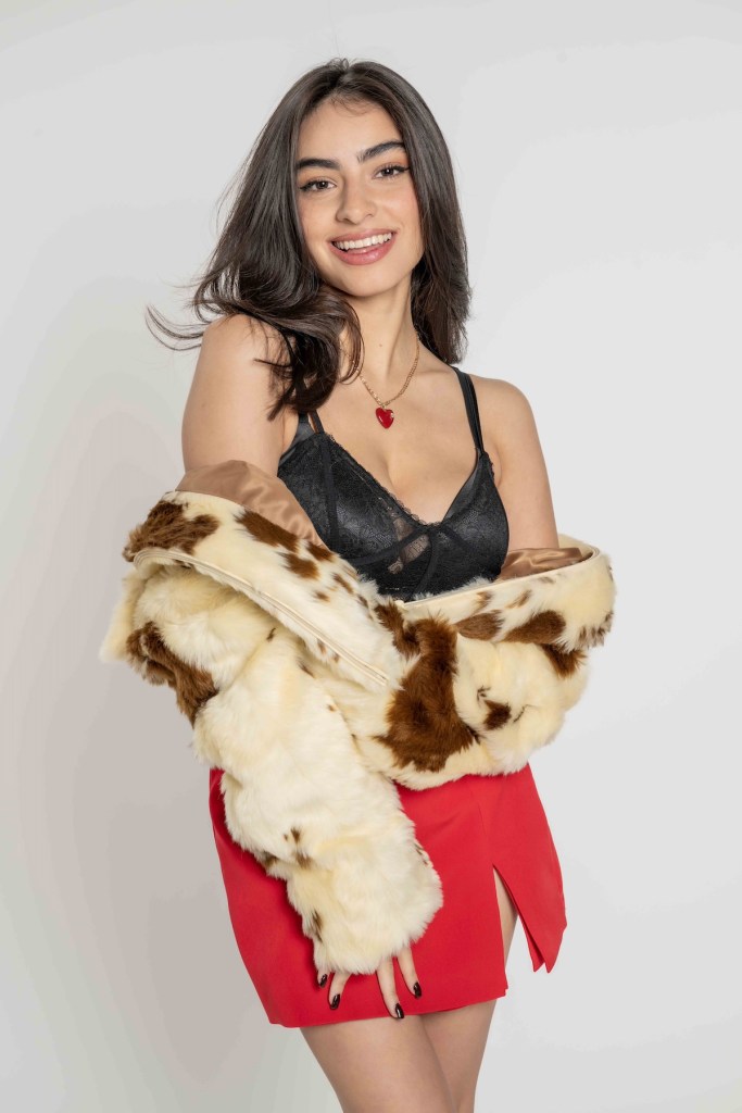 Joven posando con una blusa negra y una falda roja, sonriendo mientras sostiene una chaqueta de pelaje. Fondo blanco simple.
