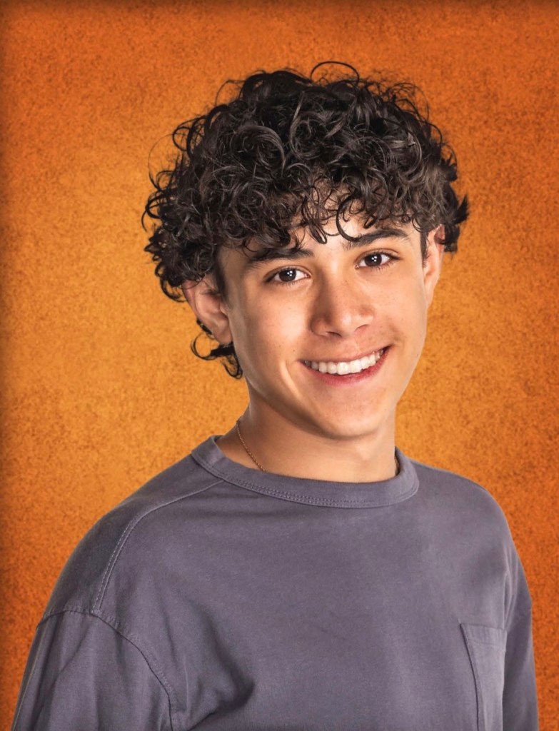 Retrato de un joven con cabello rizado y una gran sonrisa, vestido con una camiseta gris, contra un fondo naranja.