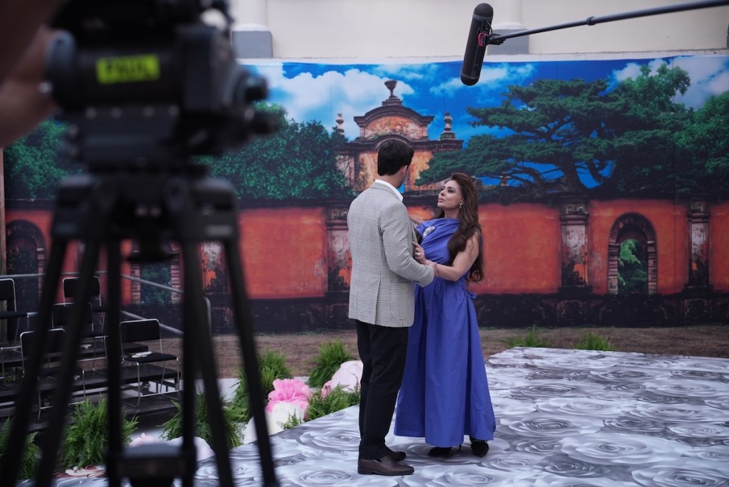 Una escena de una telenovela en grabación, donde una pareja está en primer plano frente a un fondo artístico que muestra una construcción antigua y un árbol. La mujer viste un vestido azul y el hombre lleva un traje claro, mientras un micrófono está suspendido sobre ellos.