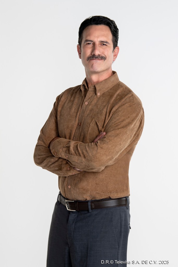Hombre de pie con camisa marrón y pantalones oscuros, sonriendo y con los brazos cruzados, contra un fondo blanco.